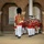 Marine Barracks Washington Evening Parade April 28, 2017 A military marching band is parading in formation along a paved walkway in front of a red brick building with arched architecture. The drum major leads the procession wearing a distinctive tall black bearskin cap and ornate red ceremonial uniform, carrying a mace. The band members behind wear matching red tunics with white trousers and white peaked caps, consistent with the United States Marine Band.
