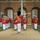 Marine Barracks Washington Evening Parade April 28, 2017 A military marching band in formal red and white dress uniforms is performing in front of a brick building with arched windows. A drum major wearing a tall black bearskin hat and red ceremonial uniform faces the band with one arm raised, directing the ensemble. The building in the background displays the date '1801', suggesting a historic military installation.
