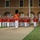 Marine Barracks Washington Evening Parade April 28, 2017 A military marching band performs in formation on a paved pathway in front of a red brick building with arched doorways. The musicians are dressed in distinctive red tunics with gold trim and white trousers, with a drum major in a tall black bearskin hat leading from the front. The setting appears to be an evening ceremonial performance at a military installation.