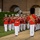 Marine Barracks Washington Evening Parade April 28, 2017 A military marching band performs in formation outdoors at night, wearing distinctive red tunics and white trousers with white peaked caps. A drum major in a tall black bearskin hat and ornate red-and-gold uniform leads the ensemble from the front. The setting appears to be a formal parade ground adjacent to a red brick building with arched windows.