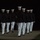 Marine Barracks Washington Evening Parade April 28, 2017 A group of military personnel in formal dress uniforms are marching in formation at night. They are wearing dark jackets with white trousers and white peaked caps, consistent with U.S. Marine Corps dress blues ceremonial attire. The figures are photographed from behind, moving away from the camera in a disciplined formation.