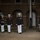 Marine Barracks Washington Evening Parade April 28, 2017 Four U.S. Marine Corps personnel in dress blue uniforms are marching in formation along a paved walkway at night. They are wearing white covers (caps), white trousers, and white gloves, with multiple medals and rank insignia visible on their uniforms. The setting appears to be a formal military ceremony at a historic brick building, likely Marine Barracks Washington or a similar installation.