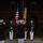 Marine Barracks Washington Evening Parade April 28, 2017 A formal military ceremony is taking place at night in front of a brick building with arched windows. Personnel in dress uniforms are rendering salutes to a color guard bearing the United States flag and what appears to be a Marine Corps battle color with campaign streamers. The scene is consistent with a U.S. Marine Corps Evening Parade or similar ceremonial event.