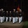 Marine Barracks Washington Evening Parade April 28, 2017 A formal military parade or ceremonial drill is taking place at night, featuring personnel in U.S. Marine Corps dress blue uniforms with white trousers and white covers. The Marines are marching in formation on a paved surface, carrying red guidon flags with gold Marine Corps insignia. The scene appears to be an evening parade ceremony, likely at a Marine Corps installation.