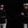 Marine Barracks Washington Evening Parade April 28, 2017 Two U.S. Marine Corps personnel in dress blue uniforms are performing a rifle drill against a black background. Both are holding wooden-stocked rifles in dynamic positions consistent with a silent drill or ceremonial performance. The dramatic lighting isolates the subjects, emphasizing the precision and formality of the display.