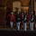 Marine Barracks Washington Evening Parade April 28, 2017 A formal military ceremony is taking place at night in front of a red brick building with arched architecture. One officer in a distinctive red dress uniform is rendering a salute while several other service members in dark blue and white dress uniforms stand at attention, with at least one holding an American flag. The scene is consistent with a U.S. Marine Corps Evening Parade or similar ceremonial event.