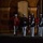 Marine Barracks Washington Evening Parade April 28, 2017 A formal military ceremony is taking place at night in front of a red brick building with arched architecture. A senior officer in a distinctive red dress uniform is rendering a salute while marching, accompanied by a color guard in dark blue and white dress uniforms holding flags. The scene appears to be a ceremonial parade or evening parade, consistent with U.S. Marine Corps formal traditions.