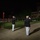 Marine Barracks Washington Evening Parade April 28, 2017 A formal military evening parade ceremony is taking place on a well-lit parade ground at night. Two officers in dress uniforms — dark jackets and white trousers — stand at salute in the foreground, facing a marching band and drill formations on the field. The setting appears to be a U.S. Marine Corps installation, consistent with a 'Evening Parade' or 'Friday Evening Parade' tradition.