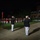 Marine Barracks Washington Evening Parade April 28, 2017 A formal military evening parade ceremony is taking place on a well-lit parade ground at night. Two officers in dark dress uniforms with white trousers and white covers are rendering salutes in the foreground, while a large military band in red and white uniforms performs in the background. Rows of uniformed Marines in dress whites are also visible in formation to the right.