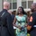 Marine Barracks Washington Evening Parade April 28, 2017 Three individuals are engaged in a friendly conversation at what appears to be a formal military event or ceremony held outdoors. Two men are wearing dark dress military uniforms, with one displaying senior enlisted rank insignia (Sergeant Major chevrons with red trim), while a woman in a floral dress stands between them smiling broadly. The setting appears to be a courtyard or garden area of an official building.