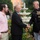 Marine Barracks Washington Evening Parade April 28, 2017 An outdoor social gathering at what appears to be a formal or semi-formal event, likely on institutional or government grounds. A U.S. Marine Corps officer in dress blues is shaking hands with a civilian man in a dark suit, while another man in a pink shirt looks on. The setting features manicured gardens, colorful flowers, and a decorative white fountain in the background.