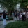 Marine Barracks Washington Evening Parade April 28, 2017 An outdoor evening gathering at what appears to be an official or formal residence, where a senior military officer in dress uniform is addressing a crowd of civilians and military personnel. The setting is a well-maintained garden courtyard with a white brick building in the background. The event appears to be a formal reception or ceremony.
