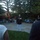 Marine Barracks Washington Evening Parade April 28, 2017 An outdoor evening military ceremony or reception taking place on a stone patio surrounded by trees and gardens. A senior military officer in dress uniform is speaking into a microphone at the center, while musicians in red uniforms perform in the background. Guests and other uniformed personnel are seated or standing around the perimeter.