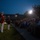 Marine Barracks Washington Evening Parade April 28, 2017 A nighttime military ceremony is taking place on a parade ground, with two U.S. Marine Corps personnel standing at attention before a large seated audience in bleachers. One Marine is dressed in the distinctive red dress uniform and is holding a French horn, indicating participation in the Marine Band. The scene is illuminated by bright stadium lights against a deep blue dusk sky, with a large brick building visible in the background.
