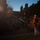 Marine Barracks Washington Evening Parade April 28, 2017 An outdoor evening ceremony featuring a military musician in a red dress uniform and white cap playing a brass instrument, likely a bugle or trumpet, before a large seated and standing audience. The scene appears to be a formal military parade or ceremonial event, possibly a sunset parade or evening colors ceremony. The low-light conditions and lens flare from artificial lighting create a dramatic atmospheric quality.
