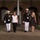 Marine Barracks Washington Evening Parade April 28, 2017 Two U.S. Marine Corps officers in dress blue uniforms escort two civilian women along a sidewalk at night in front of a brick building with arched doorways. The officers wear white covers, white gloves, and white trousers with multiple service ribbons and medals visible on their chests. The setting appears to be a formal military ceremony or evening parade event.