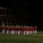 Marine Barracks Washington Evening Parade April 28, 2017 A military marching band performs an outdoor evening ceremony on a grass field, dressed in distinctive red and white uniforms. The musicians are arranged in formation while a conductor or drum major stands to the right with a raised arm and baton. A brick building with arched colonnades is visible in the background, suggesting a formal military installation.