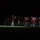 Marine Barracks Washington Evening Parade April 28, 2017 A military marching band is performing outdoors at night, illuminated by a spotlight against a dark background. The musicians are dressed in distinctive red tunics and white trousers, consistent with a formal ceremonial band uniform. A conductor or drum major stands at the front facing the ensemble, holding a baton or mace.