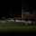 Marine Barracks Washington Evening Parade April 28, 2017 A nighttime military parade or ceremonial formation is taking place on a well-lit grass field. Rows of uniformed personnel, appearing to be U.S. Marines in dress uniforms with white trousers, are standing at attention in precise formation. Officers or inspecting officials are visible in the foreground, and a large brick building with arched colonnades is visible in the background.