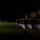 Marine Barracks Washington Evening Parade April 28, 2017 A nighttime military parade or ceremonial drill is taking place on a well-lit grass field. Two distinct formations are visible: a large column of personnel in dark dress uniforms with white trousers on the right, and a separate group in red uniforms with instruments in the background left, suggesting a marching band. The setting appears to be a formal military installation with illuminated brick buildings visible in the background.