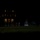 Marine Barracks Washington Evening Parade April 28, 2017 A nighttime military ceremony is taking place on a large lawn in front of a multi-story illuminated building. Personnel in dress uniforms, appearing to be U.S. Marines in red and white attire, are arranged in formation with flags visible. The scene is dramatically dark with selective lighting highlighting the ceremonial participants.