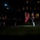 Marine Barracks Washington Evening Parade April 28, 2017 A nighttime military ceremony is taking place on a grass parade ground in front of a large illuminated building. A color guard bearer is prominently visible carrying what appears to be the United States Marine Corps flag alongside the American flag, while uniformed personnel are assembled in formation. The scene is dramatically lit with spotlights against a dark background, suggesting a formal evening parade or retreat ceremony.