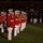 Marine Barracks Washington Evening Parade April 28, 2017 A formal military band ceremony is taking place on a grass parade ground at night, with musicians dressed in distinctive red and white dress uniforms. The band members are standing at attention in formation, holding brass instruments including trombones and French horns. In the background, a separate group of military personnel in standard dress uniforms is also standing in formation near a brick building.