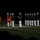 Marine Barracks Washington Evening Parade April 28, 2017 A formal USMC (United States Marine Corps) ceremonial parade or evening parade is taking place at night under artificial lighting. Marines in dress blues with white trousers and white covers are standing in formation, with two guidon bearers holding red USMC pennants labeled 'A' and 'B' from Marine Barracks Washington DC. The disciplined formation and formal attire indicate this is an official military ceremony.