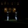 Marine Barracks Washington Evening Parade April 28, 2017 A nighttime military ceremony is taking place on a grass lawn in front of a large illuminated building. One uniformed figure stands apart playing a brass instrument (likely a bugle or trumpet) while a group of other uniformed personnel stand in formation to the right. The scene is dramatically lit with most of the surroundings in deep shadow.