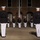 Marine Barracks Washington Evening Parade April 28, 2017 A formal military ceremony is taking place at night in front of a brick building with arched windows. Multiple service members in dress uniforms — black tunics with white trousers and white covers — are standing at attention in formation. A flag is visible on a flagpole in the background, and the building bears a partially visible date inscription reading '18__01'.