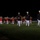 Marine Barracks Washington Evening Parade April 28, 2017 A formal military ceremonial parade is taking place at night on a well-lit grass field. Personnel in two distinct uniforms are visible: officers in dark navy/black dress uniforms with white trousers and caps in the foreground, and a marching band in red tunics with white trousers in the background. The setting appears to be an official military installation, consistent with a Evening Parade or similar ceremonial event.