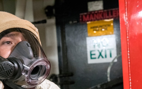 A person wearing a tan/khaki protective suit with hood and white gloves is donning or adjusting a Scott brand full-face respirator/gas mask. They are positioned near a red panel or cabinet marked 'REP 3' and 'SC', with a 'NO EXIT' sign and a 'MANKILLER' warning label visible on a dark surface behind them. The setting appears to be an industrial or military/naval environment.