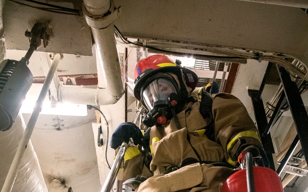A firefighter in full personal protective equipment is descending a steep metal staircase in what appears to be the interior of a ship or industrial vessel. The individual is wearing a full-face respirator mask, red helmet, tan turnout gear with yellow reflective trim, and yellow boots. A red fire extinguisher branded 'Amerex' is mounted or held near the staircase railing.