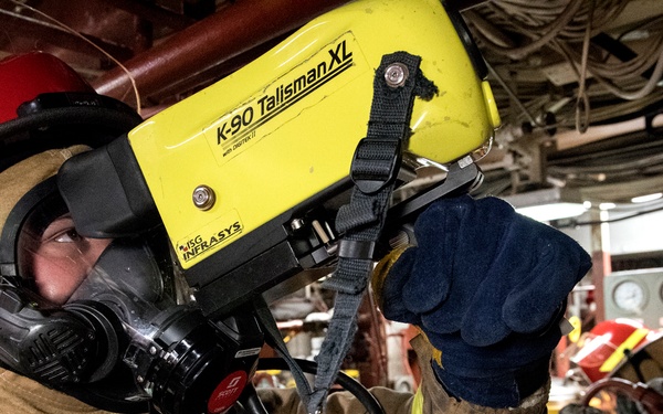 A firefighter in full personal protective equipment is operating or inspecting a yellow MSA Infrasys K-90 Talisman XL thermal imaging camera. The individual is wearing a full-face SCBA mask (Scott brand), turnout gear, red helmet, and dark blue gloves. The setting appears to be an industrial or engine room environment with visible pipes, gauges, and machinery in the background.