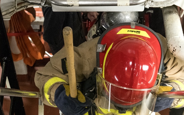A firefighter in full turnout gear is climbing down a steep metal ladder or stairway, viewed from above. The individual is wearing a red Bullard helmet with face shield, tan/yellow reflective protective gear, and carrying a wooden-handled tool, likely a fire axe or halligan bar. The setting appears to be an industrial or maritime environment based on the metal grating and red-painted structural elements.