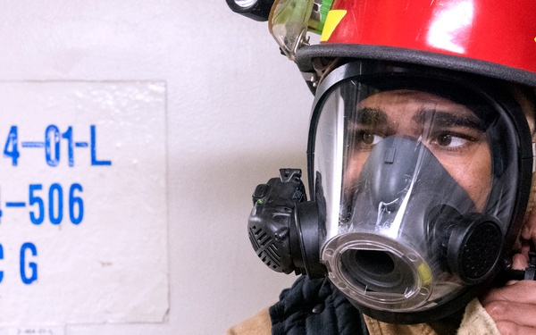 A firefighter or damage control personnel is donning a full-face respirator/SCBA mask while wearing tan firefighting gear and a red helmet with a mounted flashlight. The individual appears to be adjusting the mask straps, preparing for an emergency response or drill. A compartment identification placard on the wall behind them reads '2-464-01-L / 464-506 / CG', suggesting a naval or military vessel environment.