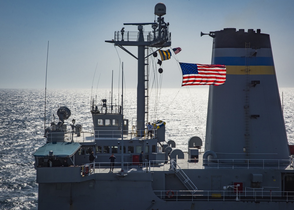 A Crew Member aboard the USNS Pecos Hoists the National Ensign During a Replenishment-at-Sea with the USS Carl Vinson