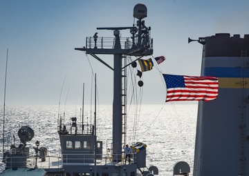 A Crew Member aboard the USNS Pecos Hoists the National Ensign During a Replenishment-at-Sea with the USS Carl Vinson