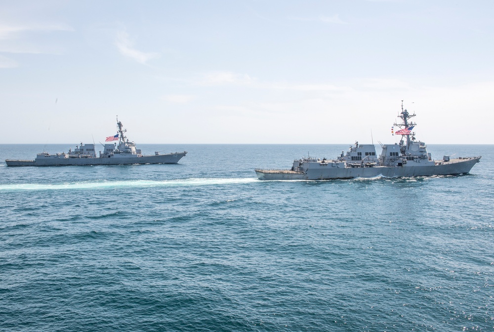 Two U.S. Navy guided-missile destroyers are underway in open ocean, sailing in close formation. Both vessels are flying American flags and appear to be Arleigh Burke-class destroyers based on their hull design and superstructure configuration. The ships are creating visible wakes as they navigate through calm to moderately choppy blue-green waters.