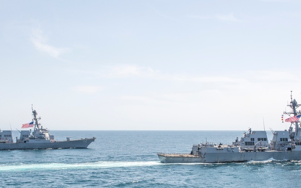 Two U.S. Navy guided-missile destroyers are underway in open ocean, sailing in close formation. Both vessels are flying American flags and appear to be Arleigh Burke-class destroyers based on their hull design and superstructure configuration. The ships are creating visible wakes as they navigate through calm to moderately choppy blue-green waters.