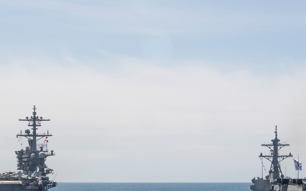 The image shows two naval vessels underway at sea in what appears to be a joint maritime operation or exercise. On the left is a large Nimitz-class aircraft carrier with aircraft visible on its flight deck, and on the right is a smaller guided-missile destroyer or frigate flying what appears to be a blue naval ensign. The vessels are sailing in relatively calm open ocean under an overcast sky.