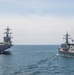 The image shows two naval vessels underway at sea in what appears to be a joint maritime operation or exercise. On the left is a large Nimitz-class aircraft carrier with aircraft visible on its flight deck, and on the right is a smaller guided-missile destroyer or frigate flying what appears to be a blue naval ensign. The vessels are sailing in relatively calm open ocean under an overcast sky.