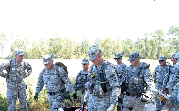 A group of U.S. Army soldiers in ACU (Army Combat Uniform) camouflage are gathered outdoors in a wooded/field training environment. Two soldiers in the center are carrying a military litter (stretcher) with medical supplies visible on it, suggesting a medical evacuation or combat casualty care training exercise. A soldier on the right appears to be taking notes or recording observations on a clipboard or notepad.