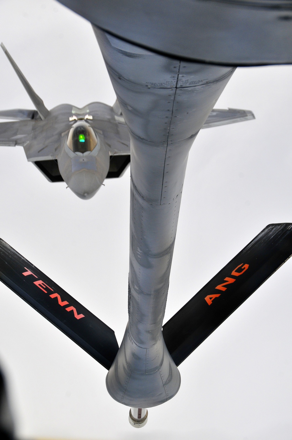 An aerial refueling operation is captured from the perspective of the tanker aircraft looking down its refueling boom toward an approaching F-22 Raptor stealth fighter. The tanker's wings are visible in the foreground, marked with 'TENN ANG' in orange lettering, identifying it as belonging to the Tennessee Air National Guard. The F-22 is positioned directly behind the boom nozzle, preparing to make contact for fuel transfer.