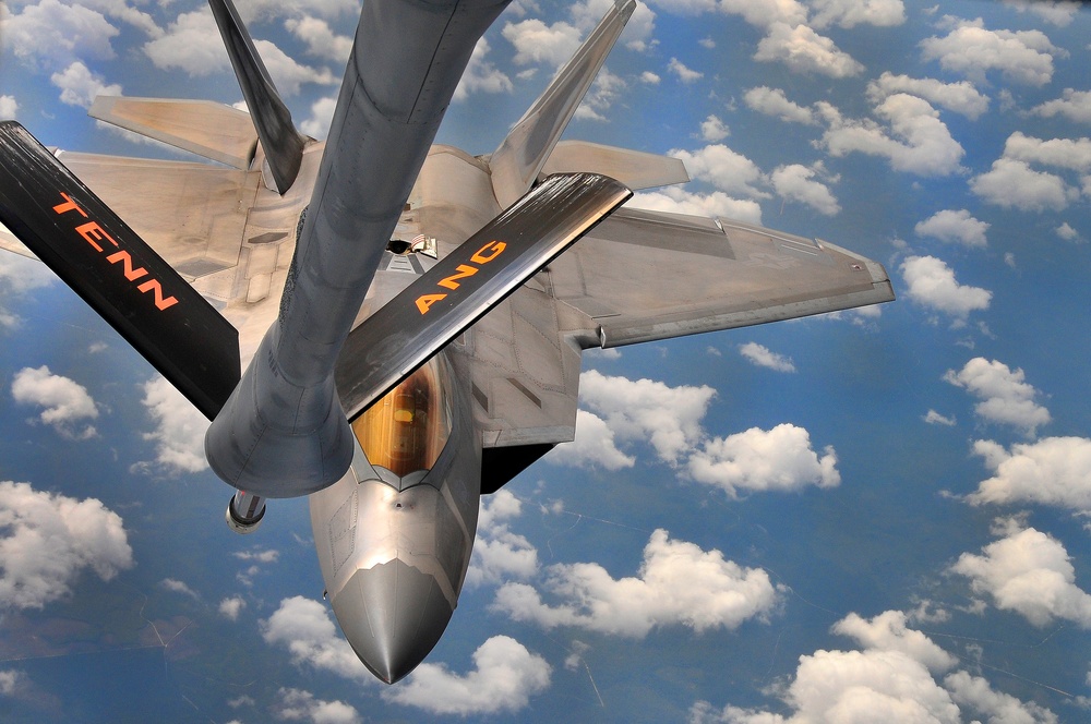 An aerial refueling operation is captured in-flight, showing an F-22 Raptor stealth fighter jet receiving fuel from a tanker aircraft via a boom refueling system. The fighter jet bears markings 'TENN' and 'ANG', indicating it belongs to the Tennessee Air National Guard. The photograph is taken from the tanker's perspective looking down at the receiver aircraft against a backdrop of scattered clouds.
