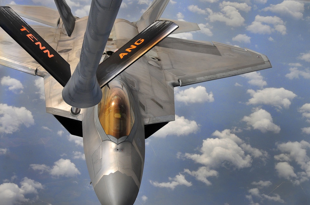 An aerial refueling operation is captured from the perspective of a tanker aircraft, showing a U.S. Air Force F-22 Raptor stealth fighter receiving fuel via a boom refueling system. The aircraft bears markings indicating it belongs to the Tennessee Air National Guard (TENN ANG), visible in orange lettering on the wings. The photo is taken at altitude with scattered cumulus clouds visible below.