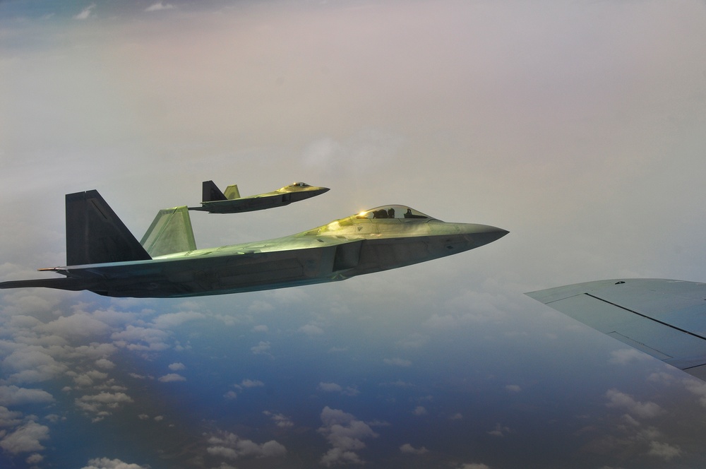 Two fifth-generation stealth fighter jets are flying in formation at high altitude above a cloud layer. The image is taken from a third aircraft, likely a tanker or transport, with its wing tip visible in the lower right corner. The aircraft are illuminated with a warm golden light, possibly from sunset or sunrise conditions.