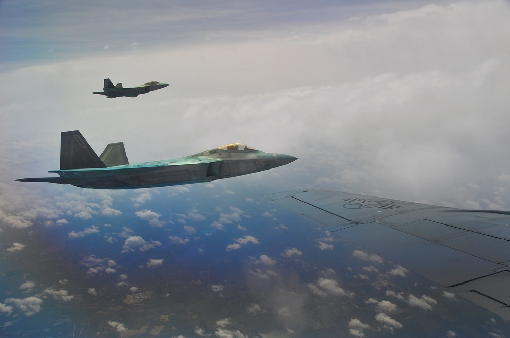 Two F-22 Raptor stealth fighter jets are flying in formation alongside a larger aircraft, from whose wing the photo is taken. The aircraft are flying at high altitude above a cloud layer with terrain visible far below. The image appears to be taken during a military escort or formation flight mission.