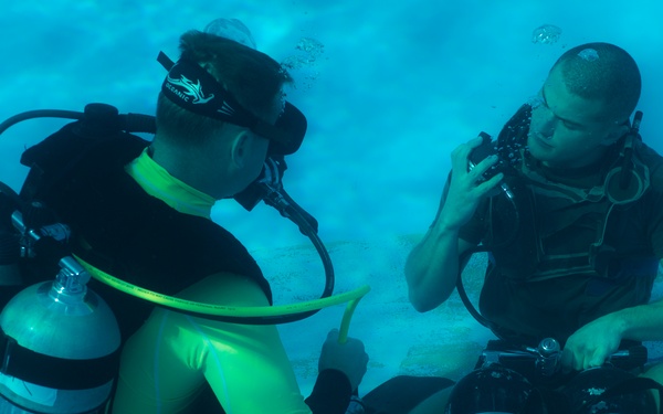 Two scuba divers are photographed underwater, appearing to be in a pool or controlled diving environment. The diver on the left is wearing a yellow-green rash guard and carrying twin scuba tanks, while the diver on the right is seated on the pool floor adjusting his mask or regulator. The image suggests a training or instructional diving session.