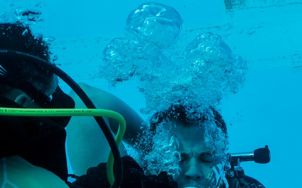 Two scuba divers are photographed underwater, appearing to be in a training or assistance scenario. One diver is exhaling air without a regulator in their mouth, producing a large cluster of bubbles rising to the surface. The other diver, wearing an Oceanic BCD and yellow hose equipment, appears to be assisting or instructing.